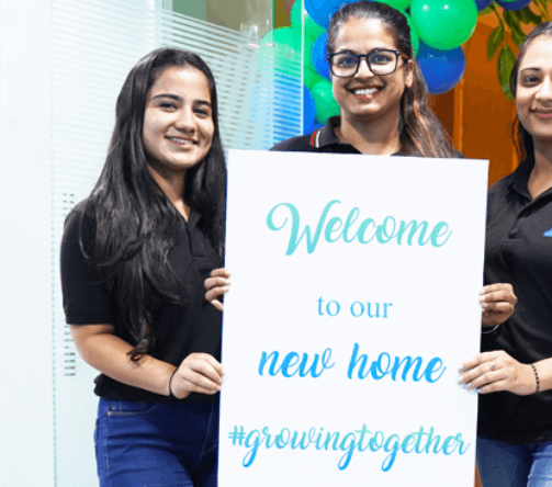 Three female employees are standing at the gate and holding a welcome banner in hand. On which is written Welcome to our new home growing together.